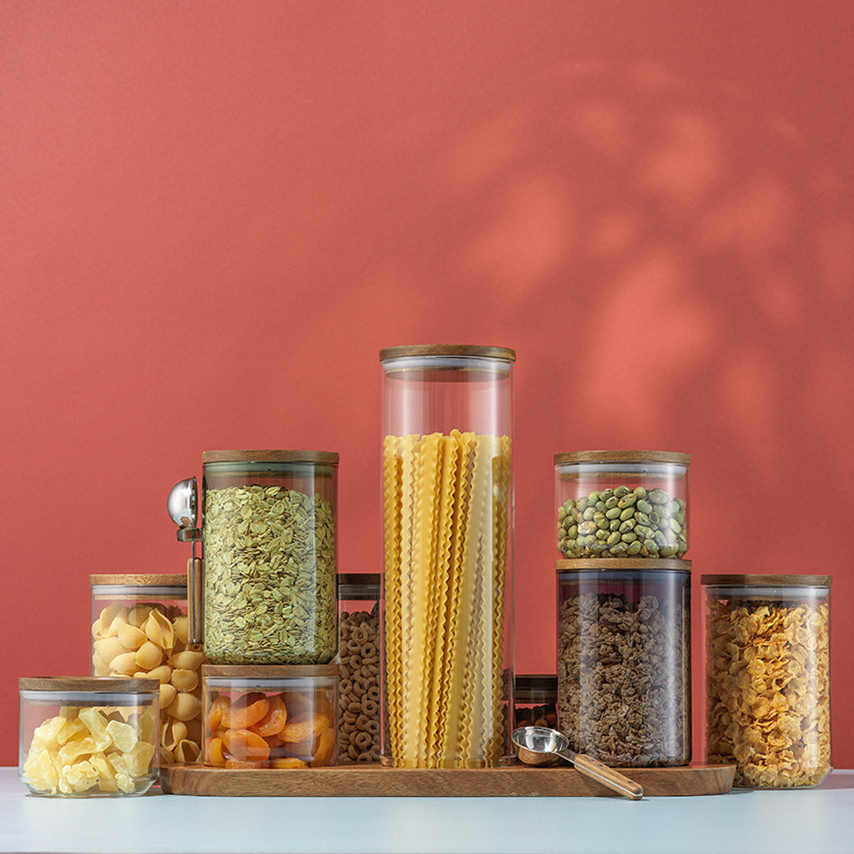 Clear glass kitchen storage containers with wooden lids filled with pasta, oats, cereal, dried fruit, and pantry essentials on a wooden tray.
