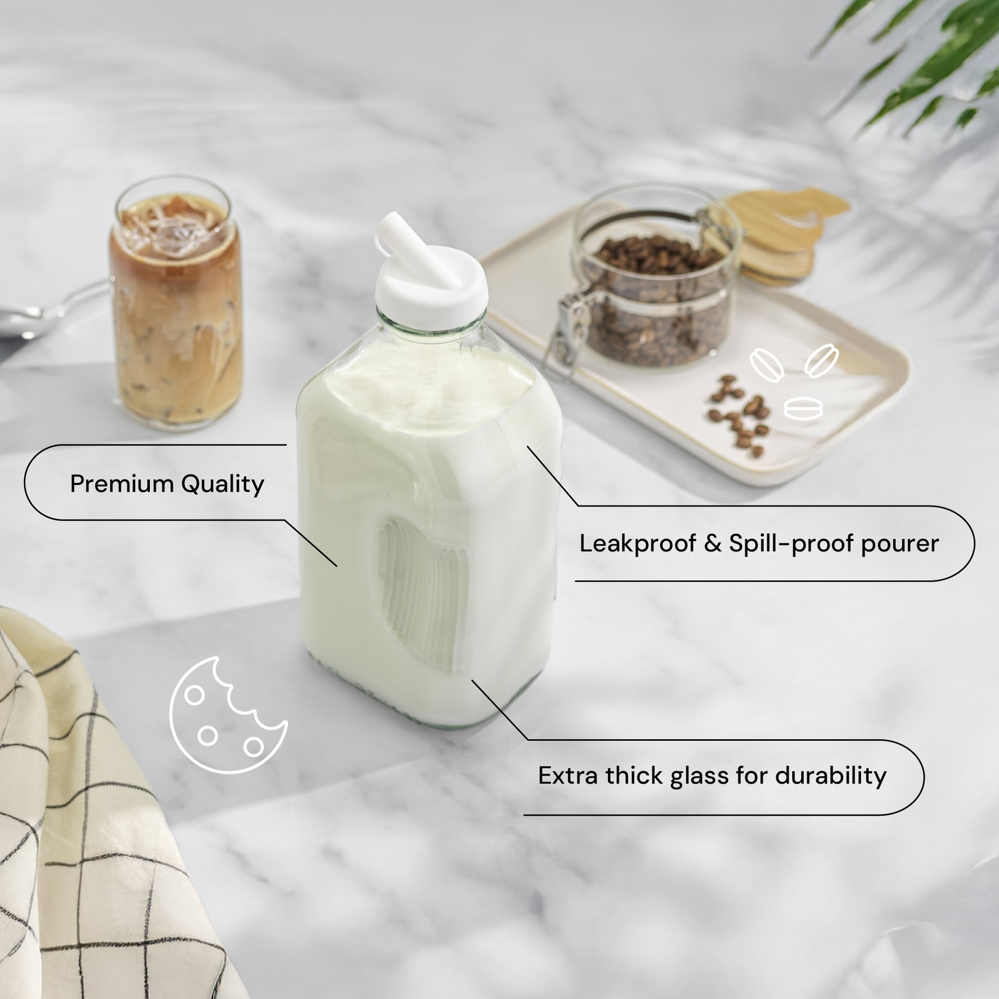 A clear milk glass bottle jug with a white pour spout, filled with milk, shown on a light marble surface with overlaid callout labels. Surrounding props include an iced coffee in a glass jar, cookies on a tray, and a folded checkered cloth.