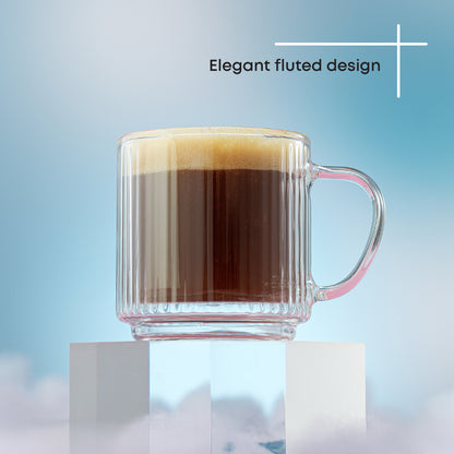 A clear ribbed glass mug filled with a dark coffee drink topped with foam, displayed on white pedestals against a soft blue gradient background with the text 'Elegant fluted design' above.