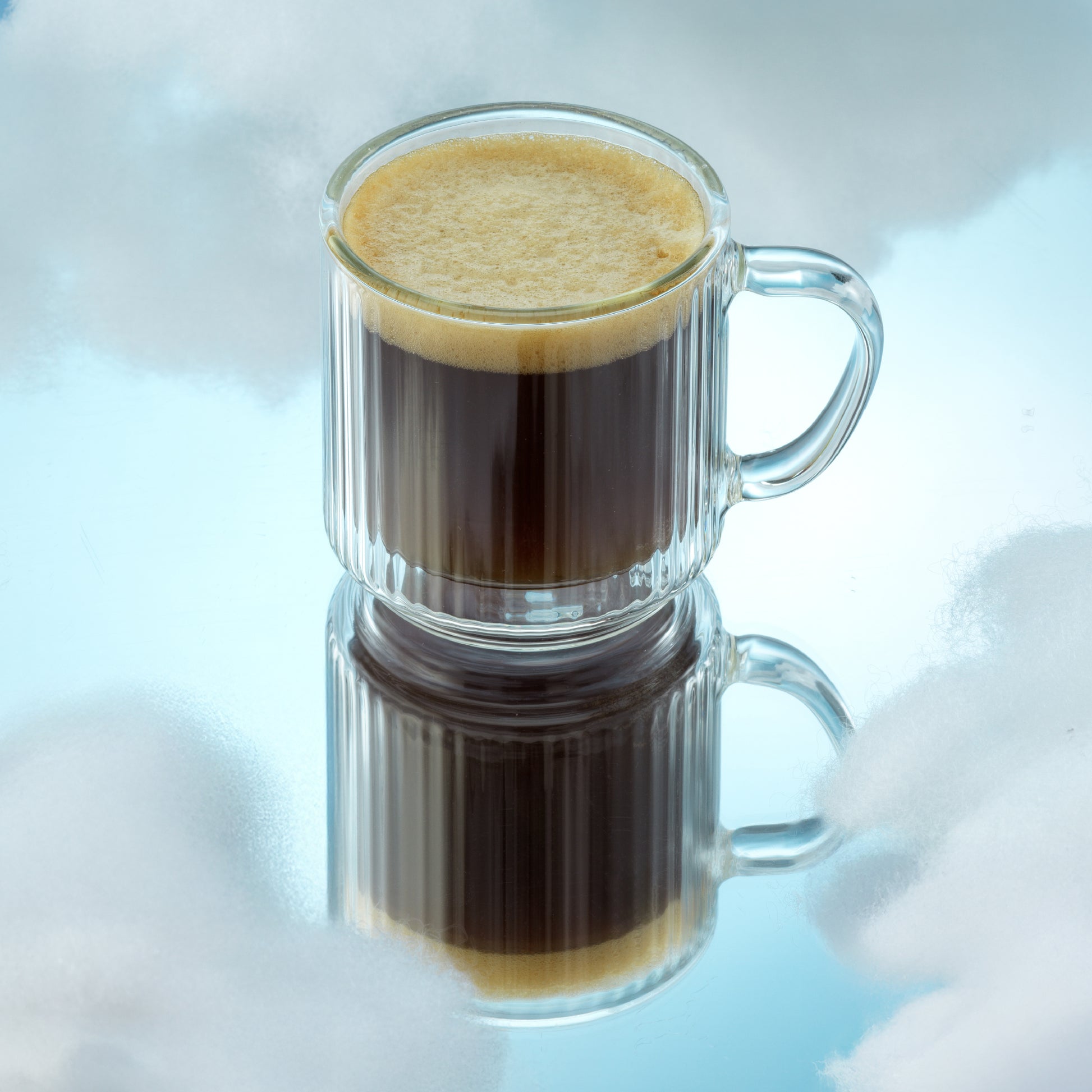A clear ribbed glass mug filled with dark coffee and topped with a layer of creamy foam, shown on a reflective light blue surface with a soft cloud-like background.