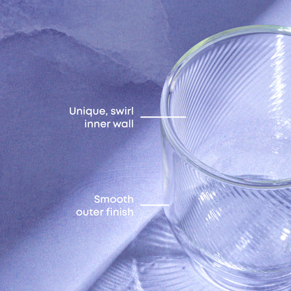 A close-up of a clear glass mug with a ribbed swirl inner wall and smooth outer finish, shown against a soft lavender-blue background with white text labels pointing to the design.