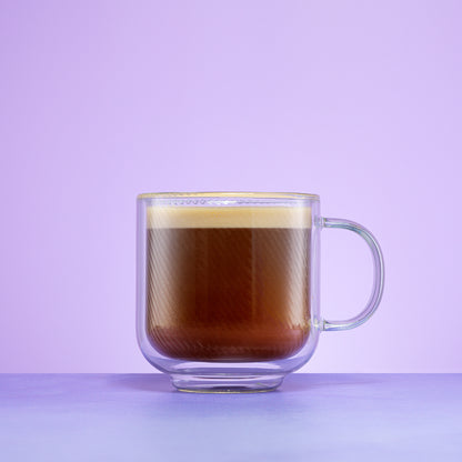 A clear double-walled glass mug filled with a layered coffee drink, set against a solid light purple background.