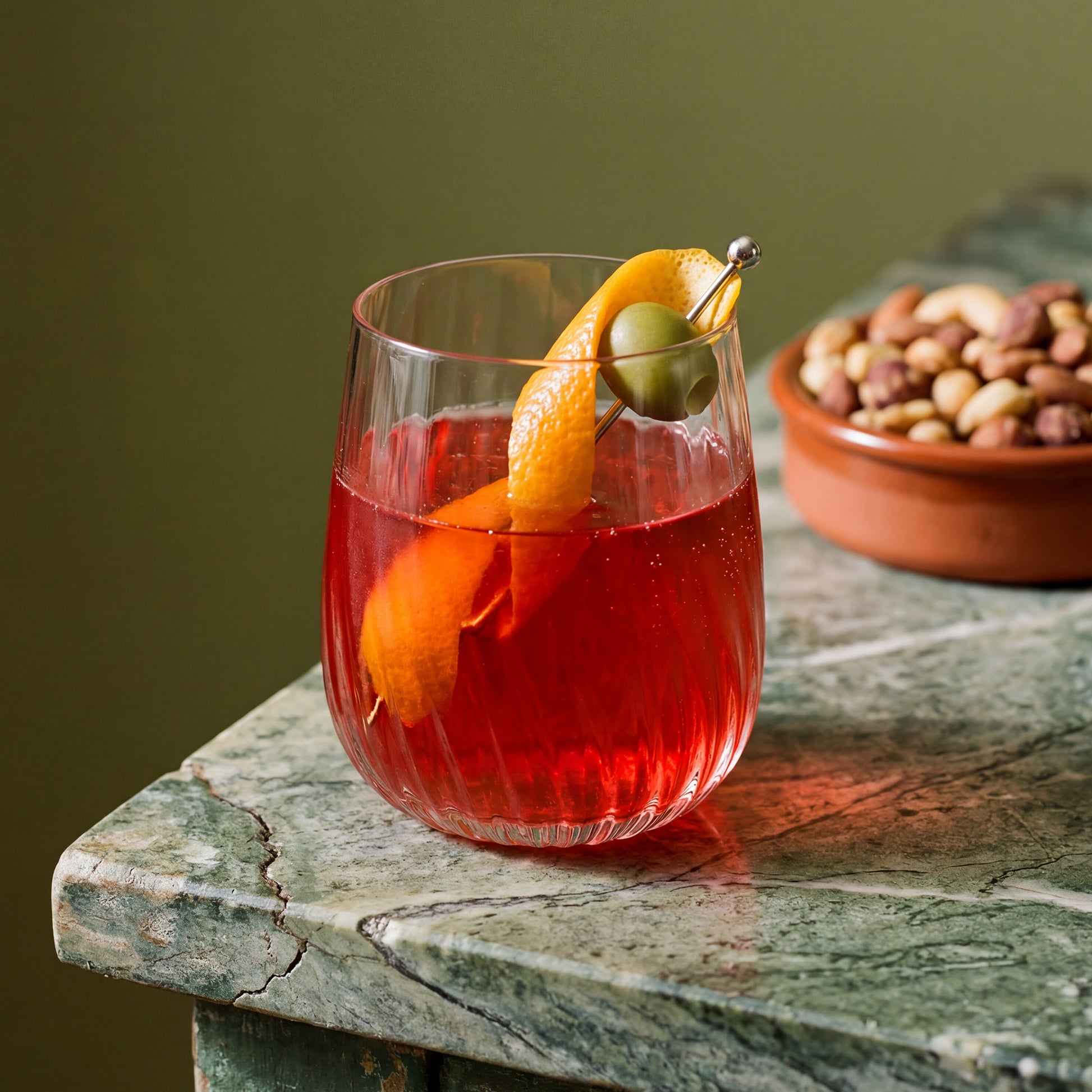 A clear glass with wavy texture filled with a red cocktail, garnished with an orange peel and a green olive on a metal skewer, set on a green marble table with a bowl of nuts blurred in the background.