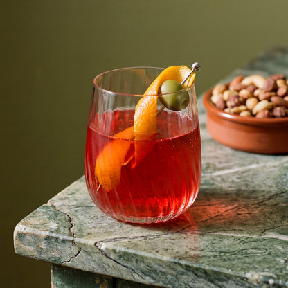 A clear glass with wavy texture filled with a red cocktail, garnished with an orange peel and a green olive on a metal skewer, set on a green marble table with a bowl of nuts blurred in the background.