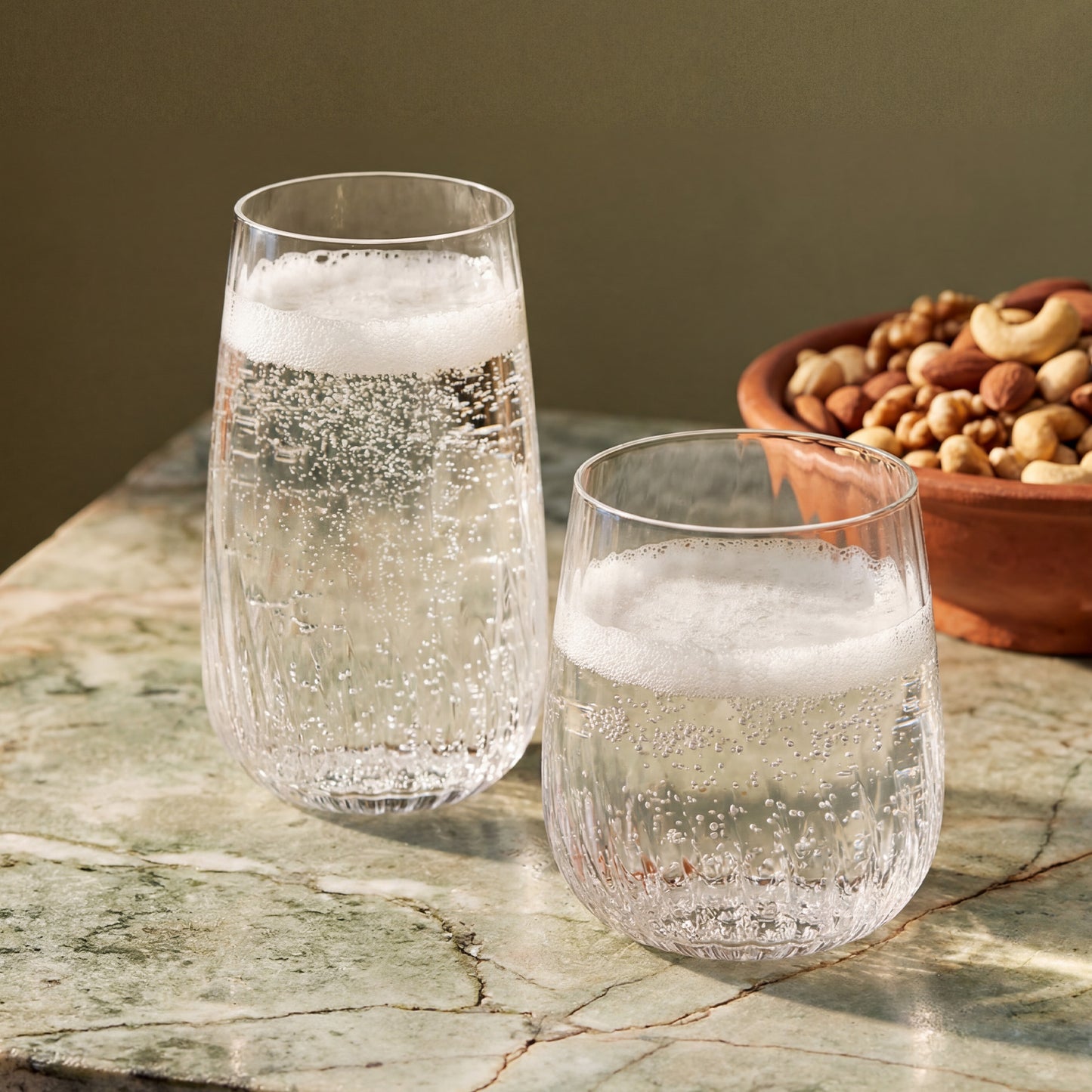 Two clear textured stemless drinking glasses filled with a bubbly carbonated drink, placed on a green marble surface with a bowl of nuts in the background.