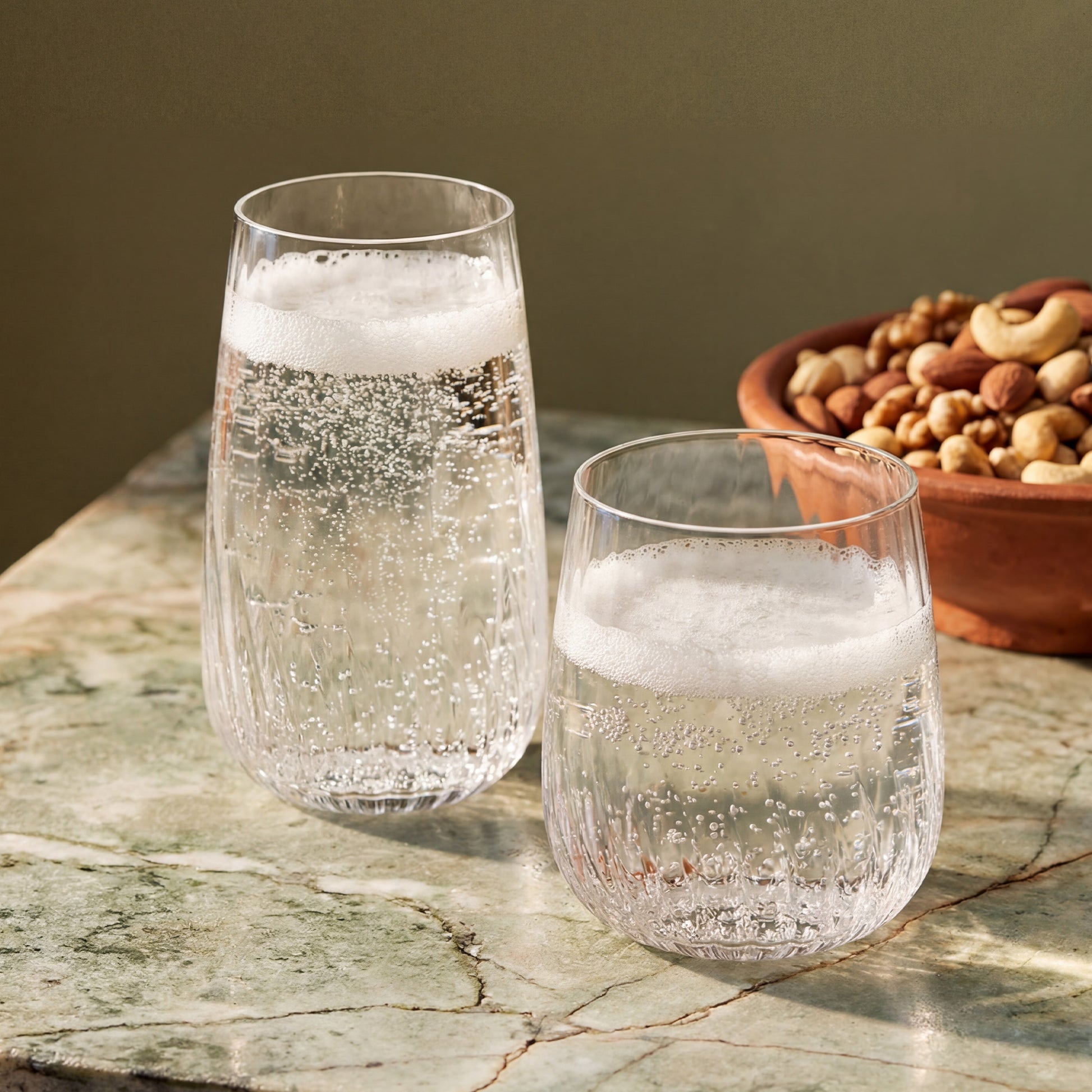 Two clear textured stemless drinking glasses filled with a bubbly carbonated drink, placed on a green marble surface with a bowl of nuts in the background.