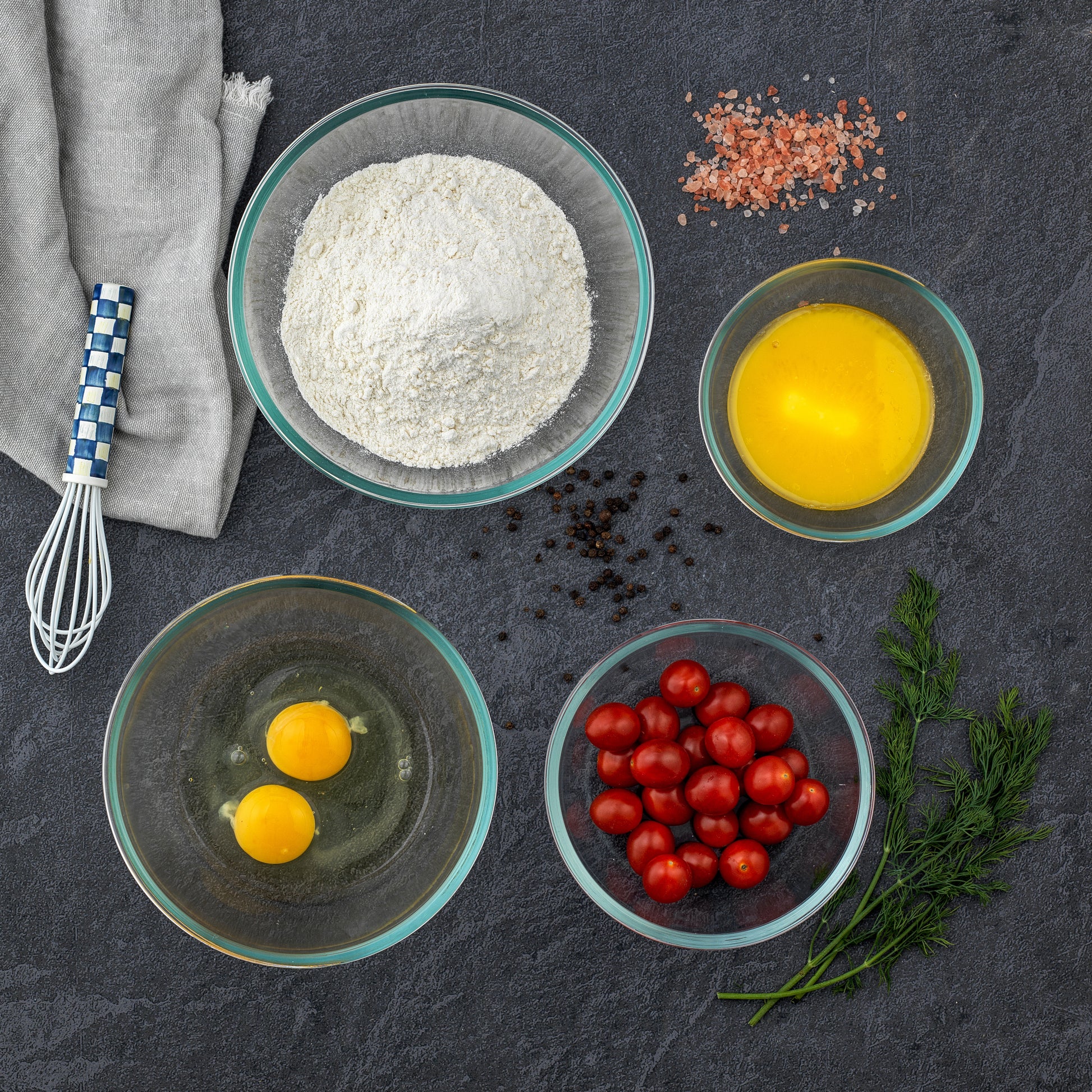 A set of three glass bowls filled with baking ingredients sits on a wooden table. From left to right, the bowls contain: white flour, two egg yolks, beaten eggs, and cherry tomatoes.